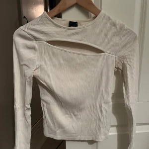 Vit ribbad topp med cut-out detalj - Snygg vit ribbad topp med lång ärm och en trendig cut-out detalj vid halsen. Perfekt för att ge din outfit en modern touch. Passar bra till både jeans och kjol.