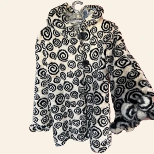 Vintage pälsjacka med spiraldesign - Unik svartvit pälsjacka med iögonfallande spiraldesign. Jackan har en stor huva och knäpps med stora knappar framtill. Insidan är fodrad med ett vackert fjärilsmönster. Perfekt för att sticka ut i mängden! Tyvär lite för liten på mig , passar S och M 