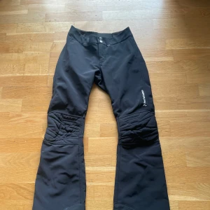 Svarta byxor från Peak Performance - Snygga svarta byxor från Peak Performance med quiltade detaljer vid knäna. Perfekta för utomhusaktiviteter med en stilren design och bekväm passform. Det finns en liten defekt på gylfen och det är en hake som saknas. 