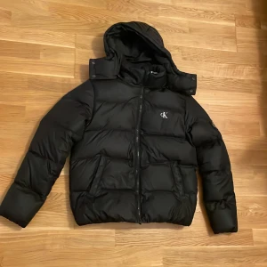 Svart dunjacka från Calvin Klein Jeans - Snygg svart dunjacka från Calvin Klein Jeans med huva och dragkedja. Jackan har en broderad logga på bröstet och är perfekt för kyligare väder.