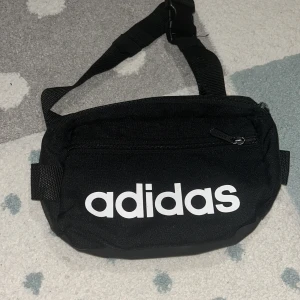 Svart magväska från Adidas - Säljer en stilren svart magväska från Adidas med justerbart bälte och dragkedja. Perfekt för att bära med sig det nödvändigaste på ett smidigt sätt. Adidas-loggan i vitt ger en sportig touch. Man kan ha den runt magen och axlarna.