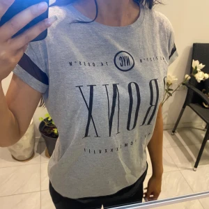 Grå t-shirt med tryck - Snygg grå t-shirt med texten 'BRONX' och 'NYC' tryckt i svart. T-shirten har korta ärmar med svarta detaljer och en rund halsringning. Perfekt för en avslappnad stil.