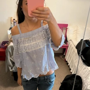 Blå- och vitrandig offshoulder-blus - Charmig blå- och vitrandig offshoulder-blus med spetsdetaljer och blommönster. Blusen har trekvartsärmar och en luftig passform, perfekt för varmare dagar.