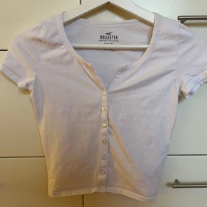 Vit kortärmad topp från Hollister storlek XS - Säljer en vit kortärmad topp från Hollister Must-Have Collection. Knappar framtill och något croppad längd. Nyskick - aldrig använd!