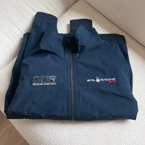 Marinblå jacka från Sail Racing - Snygg marinblå jacka från Sail Racing med dragkedja och broderad logga på bröstet. Jackan har långa ärmar och ribbade muddar vid ärmslut. Väldigt bra skick. 