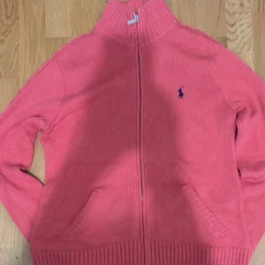Rosa kofta från Ralph Lauren - Säljer en snygg rosa kofta från Ralph Lauren med dragkedja och ribbade detaljer. Den har långa ärmar och en liten broderad logga på bröstet. Perfekt för att hålla sig varm och stilren.