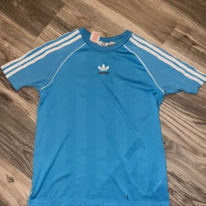 Blå t-shirt från Adidas - Säljer en blå t-shirt från Adidas med de klassiska tre ränderna på ärmarna. Tröjan har en rund hals och korta ärmar, perfekt för en sportig look. Adidas-loggan är tryckt på bröstet. Har vuxit ur den så den skulle passa bättre till nån annan😊