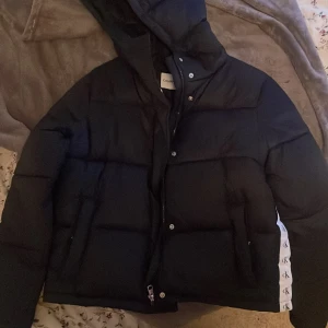 Svart pufferjacka från Calvin Klein Jeans - Snygg svart pufferjacka från Calvin Klein Jeans med huva och dragkedja. Jackan har vita detaljer med CK-logga längs sidorna och är perfekt för kyligare dagar.