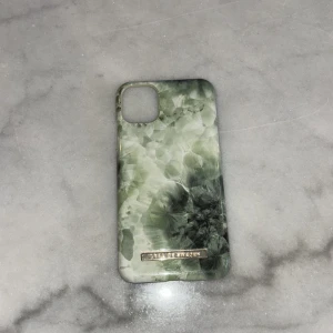 Mobilskal från Ideal of Sweden till iPhone 11 - Snyggt mobilskal från Ideal of Sweden med ett unikt mönster i gröna nyanser. Skalet har en elegant design med en marmorerad effekt och en guldig logotypdetalj. Perfekt för att ge din telefon ett stilrent utseende.
