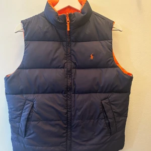 Vändbar dunväst från Ralph Lauren - Snygg vändbar dunväst från Ralph Lauren i marinblått och orange. Västen har en dragkedja framtill och två fickor på varje sida. Perfekt för att variera din stil beroende på humör och tillfälle.