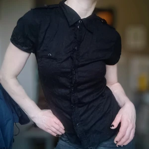 Svart kortärmad skjorta - Snygg svart kortärmad skjorta med knappar och krage. Perfekt för en stilren look. Skjortan har en ficka på bröstet och axelklaffar för extra detaljer.