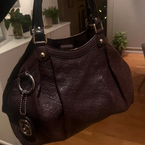 Vinröd Gucci väska med monogram - Säljer min fina Gucci-väska i mörk rött läder med deras klassiska präglade GG-monogram. Den har svarta handtag och guldfärgad hårdvara, samt en dekorativ Gucci-berlock.  Väskan har lite skavanker – insidan har några fläckar, och en kant är lite sliten. Köpt på Vestiaire, artikelnummer och ytligare bilden kan ges på begäran.