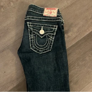 Mörkblå true religion jeans med vita sömmar - Snygga mörkblå jeans med unika broderade detaljer på bakfickorna. De har en låg midja som ger en klassisk look. Perfekta för dig som vill ha något stilrent men med en twist. Byxorna är uppsydda!