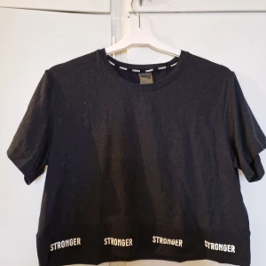 Svart t-shirt från Stronger - Snygg svart croppad t-shirt från Stronger med textdetaljer längs nederkanten. Perfekt för en avslappnad stil. Använd 2-3 gånger.