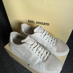 Vita sneakers från Axel Arigato - Snygga vita sneakers från Axel Arigato med stilren design. Skorna har snörning och en diskret logga på sidan. Perfekta för en minimalistisk look.