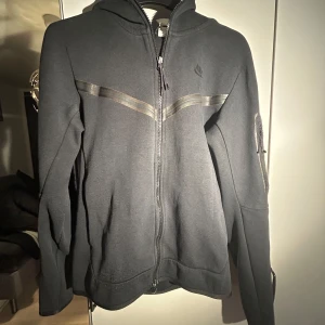Svart Nike tech hoodie, i använt skick. På baksidan är tyget lite slitet fast det går att använda, också så har en av kedjo metallen låsnat fast de är inte så märkbart - Köp för ett bra pris! En använd svart Nike tech hoodie! På baksidan är tyget lite slitet som du ser, fast det går att använda, det är också en av kedjo metallen som låsnat fast de är inte så märkbart