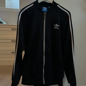 Svart träningsjacka från Adidas - Snygg svart träningsjacka från Adidas med vita ränder längs ärmarna. Jackan har dragkedja framtill och Adidas-logga på bröstet. Perfekt för sportiga aktiviteter eller en avslappnad stil.