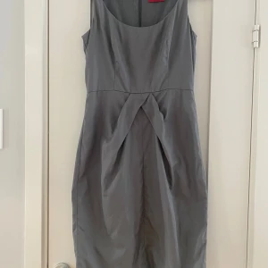 Grå armlös klänning i strl 36 från Hugo Boss. - Sparsamt använd dock finns det en liten miss i sömnaden mitt fram. I övrigt i fint begagnat skick. Klänningen har varit inlämnad hos skräddare för att korta axelbanden.  Mått Över bröstet: ca 43 cm Längd från axeln och ner: ca 93 cm 