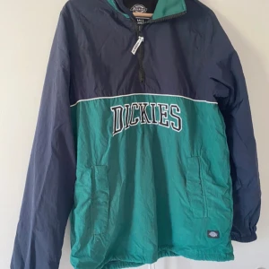 Grön och marinblå vindjacka från Dickies - Säljer en snygg och praktisk vindjacka från Dickies i storlek small. Jackan är i en cool kombination av grön och marinblå med Dickies-loggan tryckt på framsidan. Den har en dragkedja framtill och är perfekt för blåsiga dagar. Passar både till vardags och för utomhusaktiviteter.