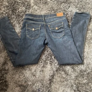 Mörkblå jeans med detaljer - Säljer ett par snygga mörkblå jeans med coola detaljer. De har en klassisk femficksdesign med två bakfickor som har knappar. Jeansen är i en rak passform och har en låg midja. Perfekta för både vardag och fest!