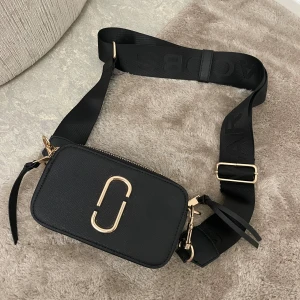 Svart axelväska med gulddetaljer - Säljer en snygg svart axelväska från Marc Jacobs. Väskan har en justerbar axelrem med märkets namn och en stilren design med gulddetaljer. Perfekt för både vardag och fest! Fick den i present så vet inte om den är äkta eller inte tyvärr, därav prisey