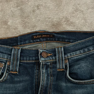 Blå jeans från Nudie Jeans - Säljer ett par snygga blå jeans från Nudie Jeans. De har en klassisk femficksdesign och är i en mörkblå tvätt med snygga slitningar. Perfekta för både vardag och fest! Skinny fit!