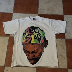 Vit t-shirt med tryck från Hellstar - Säljer en cool vit t-shirt från Hellstar med ett stort färgglatt tryck på framsidan av ett huvud med texten 'BAD BOY' i rosa och grönt på baksidan. T-shirten är i storlek S och har korta ärmar. Perfekt för en avslappnad stil!