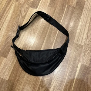 Svart midjeväska med dragkedja - Oanvänd svart midjeväska med gulddetaljer. Väskan har en justerbar rem och en praktisk dragkedja för säker förvaring. Perfekt för vardagsbruk eller resa! 