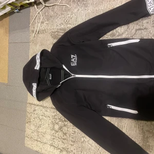 Svart hoodie från EA7 - Säljer en snygg svart hoodie från EA7 med vita detaljer. Den har en dragkedja framtill och två fickor med dragkedjor. Huvan har ett tryck med EA7-loggan. Perfekt för en casual look! Bild på kvitto finns även 