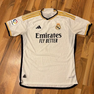 Vit Real Madrid fotbollströja - Säljer en vit Real Madrid fotbollströja från Adidas med korta ärmar. Tröjan har gula och svarta detaljer, med 'Emirates Fly Better' tryckt på framsidan. På baksidan står det 'Carvajal' och nummer 2. Perfekt för alla Real Madrid-fans!