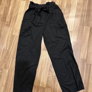 Svarta byxor med knytband - Svarta kostymbyxor med hög midja och knytband i midjan. De är i en loose passform och har en stilren design som passar perfekt till både vardag och fest. Materialet känns mjukt och bekvämt. Från Monki i storlek 40. Oanvända!!