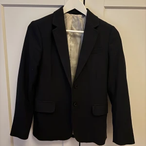 Svart kavaj i ull - Säljer en klassisk svart kavaj i ull från gant. Den har en stilren design med två knappar framtill och fickor med lock. Perfekt för både formella tillfällen och vardagsbruk. 