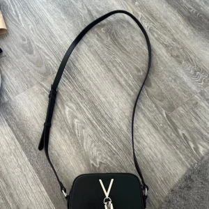 Svart axelväska med silverdetaljer - Säljer en snygg svart axelväska i skinn med en cool silverkedja och V-detalj på framsidan. Väskan har en justerbar axelrem och en praktisk dragkedja upptill. Från Mario valentino