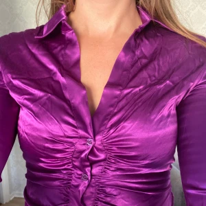 Lila satinblus - Säljer en superfin lila blus i satinmaterial. Den har en snygg v-ringning och långa ärmar med knappar vid ärmsluten. Blusen har en åtsittande passform med rynkdetaljer framtill som ger en cool look. Perfekt för fest eller en utekväll! 💜