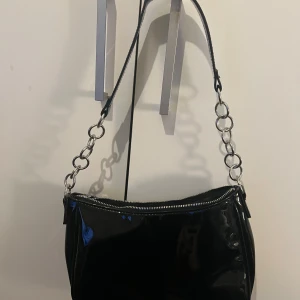Svart axelväska med kedjedetaljer - Säljer en supercool svart axelväska i syntetmaterial med glänsande finish. Väskan har en dragkedja upptill och en unik kedja som axelrem, vilket ger den en edgy look. Perfekt för en utekväll eller när du vill lägga till lite extra stil till din outfit!