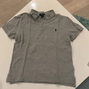 Grå pikétröja från Ralph Lauren - Säljer en stilren grå pikétröja från Ralph Lauren i mycket bra skick. Tröjan har korta ärmar och en klassisk krage med två knappar. Den är broderad med den ikoniska Ralph Lauren-loggan på bröstet. Perfekt för både vardag och fest!