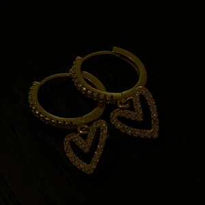 Guldörhängen med hjärtan - Snygga guldörhängen med hängande hjärtan som är täckta av glittrande stenar. Perfekta för att ge en extra touch till din outfit, oavsett om det är för en fest eller en speciell kväll. De är både eleganta och trendiga!