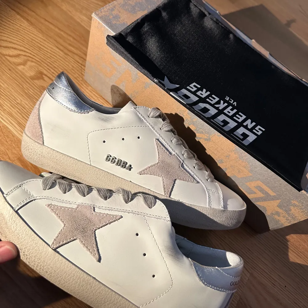 Säljer ett par snygga Golden Goose sneakers i vitt skinn med en grå stjärna på sidan. Skorna har silverfärgade detaljer på hälen och en klassisk snörning. Perfekta för både vardag och fest! De är i mycket bra skick och kommer med originalkartong.. Skor.