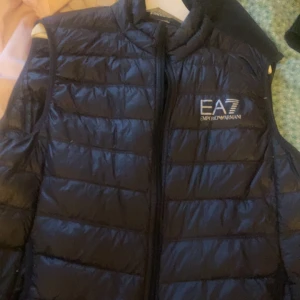 Svart dunväst från Emporio Armani - Säljer en snygg svart dunväst från Emporio Armani EA7. Västen är quiltad och har en dragkedja framtill. Perfekt för höst och vår när du vill hålla dig varm men ändå se stilren ut. Passar både till vardags och lite finare tillfällen.