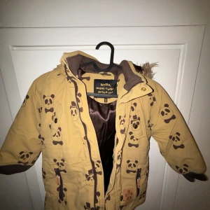 Beige jacka med pandamönster från Mini Rodini - Superfin beige jacka från Mini Rodini med gulligt pandamönster. Jackan har en mysig fuskpäls på luvan och stängs med dragkedja och knappar. Perfekt för kalla dagar och håller dig varm och stilren. Passar perfekt för höst och vinter!