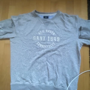 Grå sweatshirt från GANT - Säljer en grå sweatshirt från GANT i storlek L. Den har ett tryck med texten 'New Haven GANT 1949 Connecticut' på framsidan. Tröjan är i bra skick och perfekt för en avslappnad stil. Passar bra till både jeans och chinos!
