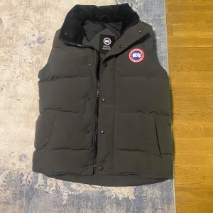 Svart dunväst från Canada Goose - Säljer en grå dunväst från Canada Goose i mycket bra skick. Västen har en quiltad design med dragkedja och knappar framtill, har ett lite varmare och mjukare material runt halsen, Perfekt för höst och vinter, håller dig varm och stilren. Passar både till vardags och lite finare tillfällen. 🖤