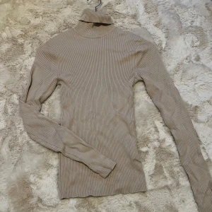 Beige ribbad polotröja - Säljer en snygg beige polotröja i ribbat material. Perfekt för höst och vinter med sin långa ärmar och mysiga polokrage. Tröjan är i mycket bra skick och passar både till vardags och lite finare tillfällen. Ett måste i garderoben för den som gillar stilrena plagg!