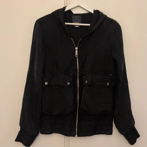 Svart jacka med huva från Diesel - Säljer en svart jacka med huva och dragkedja framtill. Den har två stora fickor med knappar på framsidan. Perfekt för höst och vår, och passar till vardags. Jackan är i bra skick och har en normal passform. 