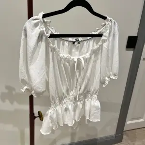 Säljer en superfin vit offshoulder blus med volangdetaljer och knyt framtill. Blusen har trekvartsärmar och en elastisk midja som ger en smickrande passform. Är ett stretchig material, så skulle säga att den passar både XS och S 💕