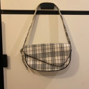 Rutig axelväska från NA-KD - Säljer en superfin rutig axelväska i svart och beige. Väskan har ett klassiskt rutmönster och en praktisk dragkedja. Perfekt för både vardag och fest! 👜