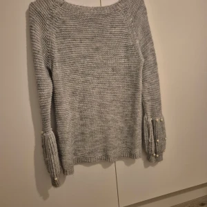 Grå stickad tröja med pärldetaljer - Mysig grå stickad tröja med långa ärmar och fina pärldetaljer vid ärmsluten. Perfekt för kyliga höst- och vinterdagar. Tröjan har en avslappnad passform och är superbekväm att ha på sig. Passar både till vardags och lite finare tillfällen.