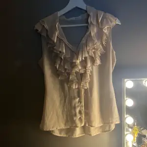 Säljer en elegant beige blus med vackra volangdetaljer framtill. Blusen har en luftig passform och är perfekt för både vardag och fest. Materialet känns mjukt och skönt mot huden. Säljer då den är för stor❤️