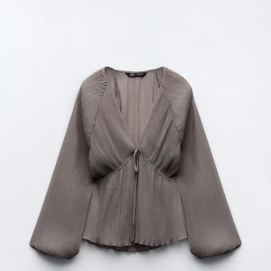 Grå plisserad blus från Zara - Snygg grå plisserad blus från Zara med långa ärmar och en knytning framtill. Perfekt för både vardag och fest. Den har en luftig passform och är gjord i ett lätt material som ger en elegant look.  Köpte den förra året i storlek XS men bara använd 2 gånger så den är helt ny ungeöför med jätte bra kvalité 