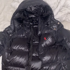 (LÄGSTA PRIS) Svart dunjacka från Ralph Lauren - Säljer en snygg svart dunjacka från Ralph Lauren. Jackan har en klassisk design med huva och dragkedja framtill. Den är perfekt för kyliga dagar och har en liten röd logga på bröstet. Passar både till vardags och lite finare tillfällen. Superbekväm och stilren! ANVÄND MAX 10 GÅNGER. KÖPTE FÖR HALVA PRISET NÄR DEN VAR PÅ REA.       KOMMER INTE GÅ NER I PRIS!!